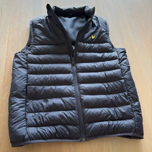 Mörkblå dunväst från Lyle & Scott - Snygg mlrkblå dunväst från Lyle & Scott med gul logga på bröstet. Västen har horisontella quiltade sömmar, hög krage och dragkedja framtill. Perfekt att slänga över en hoodie för extra värme. Klassisk och stilren design som funkar till det mesta.