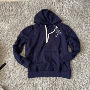 Marinblå hoodie från Polo Ralph Lauren - Snygg marinblå hoodie från Polo Ralph Lauren med vit snörning och klassisk logga på bröstet. Tröjan har huva, känguruficka och ribbade muddar vid ärmslut och nederkant. Perfekt för chill dagar och enkel att matcha med jeans eller joggers.
