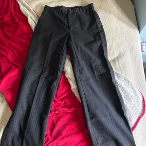 Svarta kostymbyxor med smal rand - Snygga svarta kostymbyxor med diskreta smala ränder. Klassisk rak passform och hög midja, perfekta till en dressad look. Byxorna har knapp och dragkedja framtill och är tillverkade i ett mjukt tyg som känns bekvämt hela dagen.