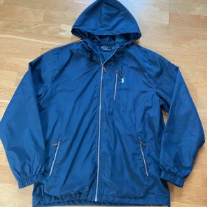 Blå vindjacka från Polo Ralph Lauren - Snygg marinblå vindjacka från Polo Ralph Lauren med huva, dragkedja framtill och tre fickor med dragkedja. Jackan har meshfoder på insidan och klassisk logga broderad på bröstet. Perfekt för dig som gillar stilren och klockren look. Kvalitet: 10/10, inga synliga defekter. Storlek L men passar M perfekt.