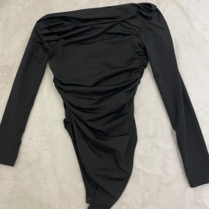 Svart offshoulder bodysuit - Snygg svart offshoulder bodysuit med långa ärmar och draperad effekt på ena sidan. Tight passform som framhäver siluetten och ger en trendig look. Perfekt att styla med jeans eller kjol för en cool outfit.
