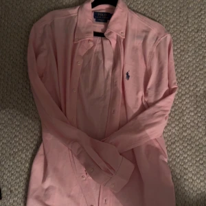 Rosa skjorta från Polo Ralph Lauren - Snygg rosa skjorta från Polo Ralph Lauren med klassisk krage och knappar framtill. Skjortan har lång ärm och ikoniska broderade loggan i mörkblått på bröstet. Perfekt för dig som gillar stilrena och tidlösa plagg.