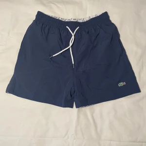 Lacoste badshorts  - Snygga marinblå badshorts från Lacoste med klassisk krokodillogga på benet. Shortsen har elastisk midja med vit snörning och en bakficka med lock. Perfekta för stranden eller poolen, riktigt clean look och skön passform.