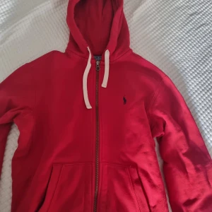 Röd hoodie från Polo Ralph Lauren - Säljer en röd hoodie från Polo Ralph Lauren i storlek M. Tröjan har vit snörning i huvan, dragkedja framtill och två stora fickor. Klassisk Polo-logga broderad på bröstet. Se gärna mina andra annonser och skapa bundles. Se bilder för mått. 