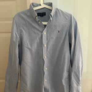 Ljusblå skjorta från Ralph Lauren - Klassisk ljusblå skjorta från Ralph Lauren med button-down krage och broderad logga på bröstet. Skjortan har långa ärmar och vita knappar framtill. Perfekt för en stilren och avslappnad look.