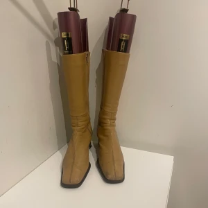 Boots  - Högklackade stövlar/boots. Väldigt sköna att gå i. Märket är okänt. Slitet skick, se klacken på sista bilden. Detta är dock inget som märks tydligt när skorna används eller påverkar bekvämligheten. Färgen är brunbeige