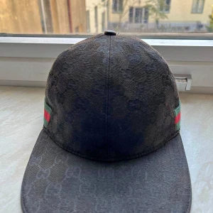 Svart Gucci keps med GG-mönster - Snygg svart keps från Gucci med klassiskt GG-monogram över hela kepsen och grönt/rött band på sidorna. Justerbar rem baktill i skinn. Perfekt accessoar för dig som vill sticka ut med lyxig streetstyle.