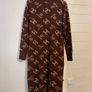 Brun långärmad  klänning - Säljer en brun långklänning med beige logomönster över hela klänningen. Den har långärmade ribbade ärmslut och är stickad i ett mjukt material. Perfekt för dig som vill ha en statement piece med lyxig känsla och snygg passform.