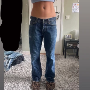 Blå jeans från Evisu, storlek 30 - Säljer ett par blå jeans från Evisu i klassisk straight fit med coola vita logobroderier på bakfickorna. Jeansen är tillverkade i 100% bomull och har låg midja. Perfekt för dig som gillar streetwear och vill sticka ut med snygga detaljer.