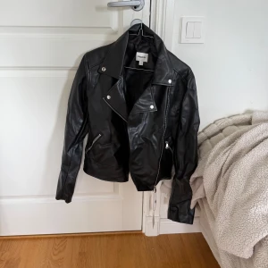 Svart skinnjacka från Kappahl - Snygg svart skinnjacka från Kappahl i bikerstil med silvriga dragkedjor och knappar. Jackan har klassisk krage, långa ärmar och coola detaljer som ger en edgy vibe. Perfekt för dig som vill ha en tidlös och trendig look.