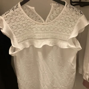 Vit blus med spets och volang - Superfin vit blus från Mango med detaljerad spets upptill och volang längs axlarna. Blusen är ärmlös och har en rund halsringning med vackert broderat mönster. Perfekt för dig som gillar romantiska och luftiga plagg.