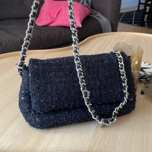 Svart axelväska Becksöndergaard - Säljer en svart axelväska i tweedliknande tyg med små glittrande paljetter i olika färger. Väskan har 2 defekter där av priset: tyget har släppt vid knappen på framsidan, men går fortfarande att använda. Sedan har kedjan släppt på ena sidan. Jag brukar använda väskan som en clutch🥰 Nypris 850kr