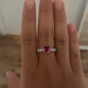 Silverring med hjärtformad röd sten - Snygg silverfärgad ring med en stor hjärtformad röd sten i mitten, omgiven av små glittrande stenar längs bandet. Perfekt för dig som gillar statement-smycken och vill ha något som verkligen sticker ut. Ringen har en romantisk vibe och passar dig som älskar detaljer.äkta silver och aldrig använt.
