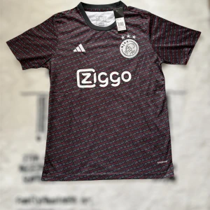 Ajax x Adidas svart fotbollströja S - Ajax fotbollströja från Adidas i svart med röd och grå grafisk design. Tröjan har korta ärmar, rund hals och Ajax klubbmärke broderat på bröstet. Materialet är lätt och andas, perfekt för match eller träning. Ziggo-logga på framsidan.