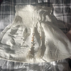 Vit kortkjol med smock och pärldetaljer - Supersöt vit kortkjol med smockad midja och dekorativa knytband med pärlor och tofsar. Kjolen har en lätt volangig form och är gjord i ett mjukt bomullstyg. Perfekt för varma dagar när du vill känna dig både chill och stylish.