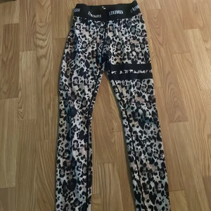 Leopardmönstrade leggings från Stronger - Snygga leopardmönstrade leggings från Stronger i storlek S. De har elastisk midja med logga och är i stretchigt material som passar perfekt till träning. Färgerna går i svart, beige och brunt med coola svarta detaljer på benen. 