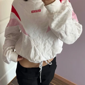 Vit och röd croppad Levi's tröja XS - Säljer en croppad sweatshirt från Levi's i storlek XS. Tröjan är vit med röda och rosa detaljer, har lång ärm och snörning med logga i nederkant. Snyggt mönster framtill och loose passform, perfekt för dig som gillar streetstyle.