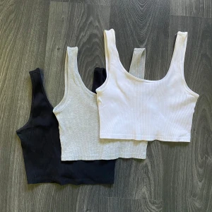 3-pack ribbade croppade linnetoppar - Tre croppade linnetoppar i ribbad bomull, perfekta för layering eller chill stil. Färgerna är svart, ljusgrå och vit, alla med breda axelband och rundad hals. Snygga att matcha med jeans eller shorts för en enkel look. 