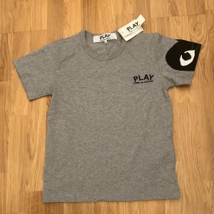 t-shirt Comme des Garçons Play - Grå t-shirt från Comme des Garçons Play med svart logga på bröstet och en stor svartvit detalj på ena ärmen. Klassisk rund halsringning och korta ärmar. Skön och stilren, perfekt för dig som gillar streetwear och minimalistisk design.