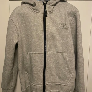 CP Company grå zip - Hej, jag säljer en CP company zip som är grå oanvänt skick. Lappen sitter fortfarande kvar på insidan. Det ända dåliga är att en liten bit av texten på högra glasögat är avflagnat från gången jag använde den.