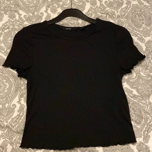 Svart ribbad topp från Vero Moda - Svart ribbad topp från Vero Moda med korta ärmar och vågiga kanter vid ärmslut och nederkant. Enkel och stilren design som funkar till många olika looks. Mjuk och stretchig känsla, perfekt för dig som gillar basic med en twist.