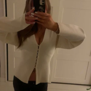 Beige kofta med v-ringning och knappar - Säljer en stilren beige kofta med v-ringad hals och knappar framtill. Koftan har långärmade, lite vidare ärmar och är gjord i ett mjukt material som känns skönt mot huden. Perfekt för dig som gillar enkel och clean stil. En av knapparnas spänne har lossats, men är tillbaka knutet, samt en liten fläck , kom privat för mer bilder 