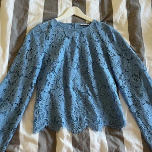 Blommig blå spetsblus från Bubbleroom - Supersöt blå blus från Bubbleroom i genombruten spets med blommigt mönster. Långärmad modell med rund halsringning och knapp i nacken. Perfekt för dig som vill ha en romantisk och trendig look med fina detaljer.