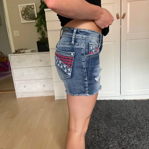 Blå jeansshorts med stjärndetaljer - Snygga blå jeansshorts med slitna detaljer och fransig kant. Bakfickorna har broderade stjärnor och röda ränder som ger en cool, amerikansk vibe. Shortsen är midwaist och har klassisk femficksdesign. Perfekta för sommaren och festivaler!