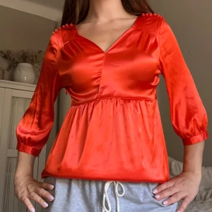 Röd satinblus med v-ringning - Snygg röd blus i glansigt satinmaterial med v-ringad hals och puffiga trekvartsärmar. Axelpartiet har små dekorativa knappar och blusen har en lätt volang under bysten för en chill och feminin look. Perfekt för dig som gillar färg och vill sticka ut.