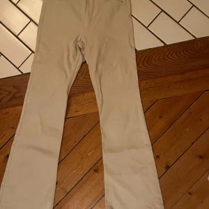 Beiga byxor från H&M - Säljer ett par stilrena beiga byxor från H&M i bootcut modell. Byxorna har klassisk femficksdesign, bälteshällor och silverfärgade knappar. Perfekta för en clean och enkel look. Materialet är mjukt och känns skönt mot huden.