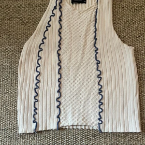 Beige stickad linnetopp från Cider - Säljer en beige stickad linnetopp från Cider i storlek M. Toppen har ribbad struktur på sidorna och ett hålmönstrat parti i mitten. Två vågiga blå detaljer löper vertikalt framtill och ger en cool kontrast. Perfekt för dig som gillar unika detaljer och skön passform.