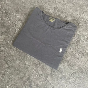 Mörkgrå t-shirt från Polo Ralph Lauren - Snygg mörkgrå t-shirt från Polo Ralph Lauren i klassisk modell med korta ärmar och rund hals. T-shirten har en liten broderad logga på bröstet och är tillverkad i mjuk bomull, perfekt för en chill och stilren look. 