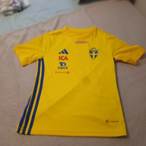 Svenska landslagets fotbollströja Adidas - Gul fotbollströja från Adidas med blå detaljer och svenska landslagets emblem på bröstet. Tröjan har korta ärmar, rund hals och tryckta sponsorer som ICA, OBOS, Swedbank och Bauhaus. Materialet är lätt och andas, perfekt för träning eller match.
