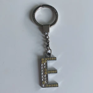 Nyckelring med glittrig bokstav E - Snygg nyckelring i silverfärgad metall med en stor bokstav E dekorerad med glittrande stenar i gult och vitt. Perfekt accessoar för att göra dina nycklar extra personliga och blingiga.