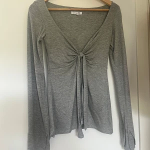 Grå långärmad topp med knyt från Design Si - Fin grå långärmad topp från Design Si med djup v-ringning och knyt framtill. Tunn och mjuk i materialet. Inte använd 