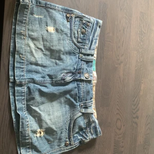 Blå jeanskjol från Miss Denim - Säljer en kort blå jeanskjol från Miss Denim med slitna detaljer och coola fickor med nitar och broderade sömmar i rosa och orange. Kjolen har klassiska jeansknappar och bälteshällor. 