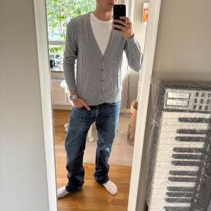 Grå kofta med knappar - Snygg grå kofta med v-ringning och knappar framtill. Modellen är klassisk och passar perfekt över en t-shirt. Koftan har långa ärmar och ribbade muddar vid ärmslut och nederkant. Materialet känns mjukt och lätt, troligen bomull eller en bomullsblandning.