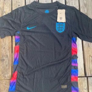England svart fotbollströja Nike - Snygg svart England fotbollströja från Nike med blått logotryck och coola färgdetaljer i rött, blått och lila längs sidorna. Kortärmad modell i lätt och ventilerande material, perfekt för match eller träning. Modern design med diskreta ränder i tyget. säljer så snabbt som möjligt så tveka inte att komma med bud. Passar även storlek M