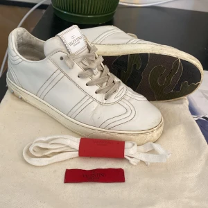 Valentino Garavani vita sneakers - Säljer ett par vita Valentino Garavani sneakers med c nitar på sidan . Sulan har ett unikt camouflage-mönster i grönt och svart. Med extra vita skosnören och röd etikett. Perfekt för dig som gillar exklusiv streetstyle. Saknar två stycken taggar på baksidan. Annars bra skick.  Skriv gärna 😊