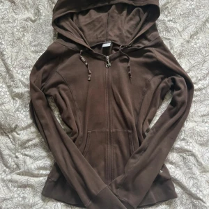 Brun hoodie från DEHA med dragkedja - Säljer en brun hoodie från DEHA i storlek L. Tröjan har huva med snörning, dragkedja framtill och en stor ficka på magen. Mjuk bomullskänsla och diskret logga på fickan. Perfekt för chill dagar eller när du vill vara extra bekväm.