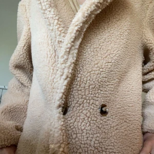 Beige teddyjacka från EillyBazar - Supermysig beige teddyjacka från EillyBazar med oversized passform och stora knappar framtill. Jackan har lång ärm och är gjord i fluffigt syntetmaterial som ger en cool och trendig look. Perfekt för kalla dagar när du vill vara både varm och snygg.