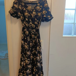 Mörkblå blommig långklänning med volang - Supersöt långklänning i mörkblått med blommigt mönster och volangiga korta ärmar. Klänningen har v-ringning, knytband i midjan och ett flowigt fall. Perfekt för dig som gillar romantisk stil och vill sticka ut lite extra.