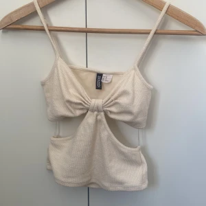 Beige croppad topp från H&M Divided - Snygg beige croppad topp från H&M Divided med smala axelband och cool cut-out detalj framtill. Toppen har en knytning i mitten som ger en unik look och är tillverkad i mjukt ribbat material. Säljer då den är alldeles för liten för mig, skulle säga att tröjan passar någon som gillar tajta tröjor eller som är petite. 