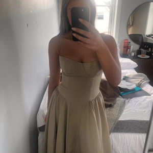 Beige långklänning med smala axelband - Säljer en stilren beige långklänning med smala axelband och markerad midja. Klänningen har en feminin siluett med lätt utställd kjol och bystdetaljer. Perfekt för sommarens tillfällen när du vill känna dig extra fin. Med den stora kupan 