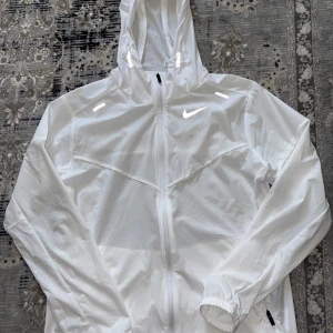 Vit vindjacka från Nike - Snygg vit vindjacka från Nike med huva och dragkedja hela vägen. Jackan har två fickor med dragkedja och reflekterande detaljer på axlarna. Perfekt för blåsiga dagar och sportiga outfits.