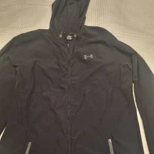 Svart vindjacka med huva från Under Armour - Svart vindjacka från Under Armour med huva och dragkedja. Jackan har två fickor med dragkedja och en diskret logga på bröstet.