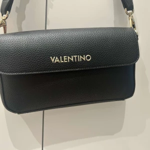 Svart axelväska från Valentino - Snygg svart axelväska från Valentino med guldiga detaljer och logotyp framtill. Väskan har en bred axelrem med stora bokstäver i guld och är tillverkad i ett strukturerat skinnliknande material. Perfekt för att lyfta din outfit.