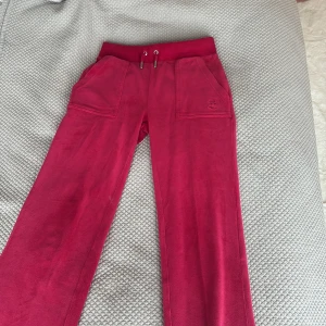 Rosa mjukisbyxor från Juicy Couture - Säljer ett par klassiska rosa mjukisbyxor från Juicy Couture i supermjuk velour. Byxorna har dragsko i midjan, stora fickor fram och bak samt broderad logga på ena benet. Perfekta för en avslappnad och trendig look.