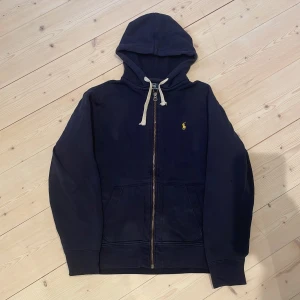 Mörkblå hoodie från Polo Ralph Lauren - Säljer en mörkblå hoodie från Polo Ralph Lauren med dragkedja och vit snörning i huvan. Klassisk gul broderad logga på bröstet och två fickor framtill. Perfekt för en avslappnad stil. Storlek L men passar M. Skick 9/10, det är bara att skriva om ni undrar något!