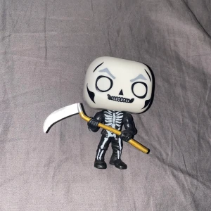 Fortnite gubbe funko pop - Bilden visar en Funko Pop!-figur föreställande ett skelett med lie, men inget bokomslag eller bokinformation syns. Kan vara relaterad till populärkultur eller samlarobjekt.